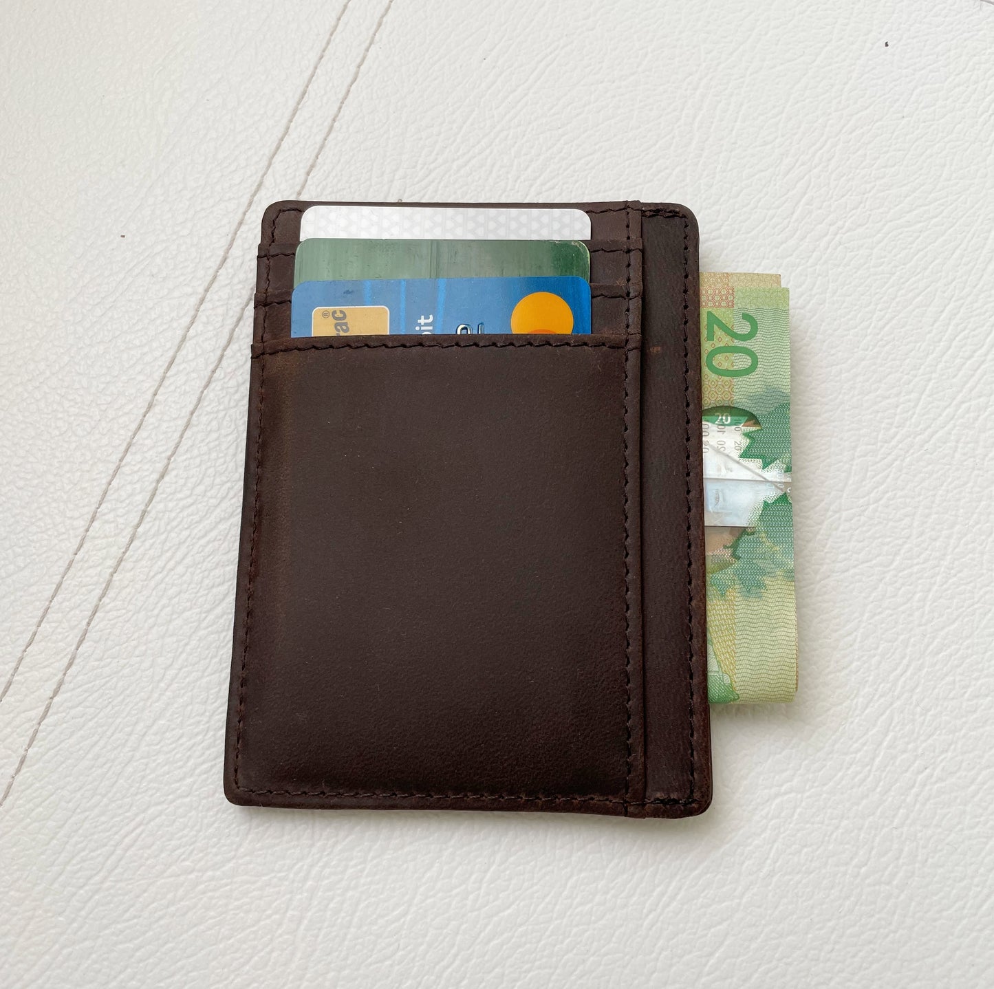 A dark brown leather minimalist cardholder wallet shown closed on a light textured surface, with cards and a 20 Australian dollar banknote partially visible in the slots.