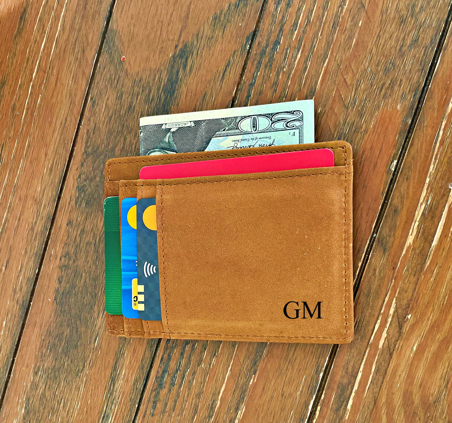 A tan leather bifold wallet with card slots and a dollar bill partially visible, placed on a rustic wooden surface. The wallet is personalized with the initials 'GM' in black on the front.