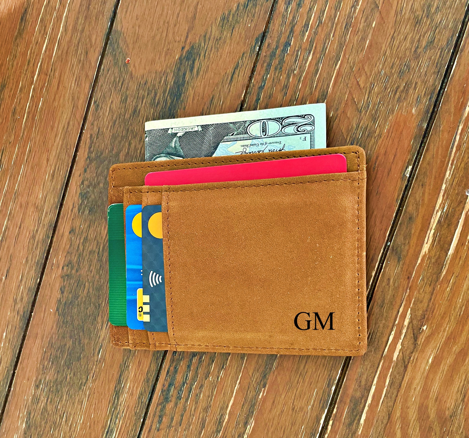 A tan leather bifold wallet with card slots and a dollar bill partially visible, placed on a rustic wooden surface. The wallet is personalized with the initials 'GM' in black on the front.