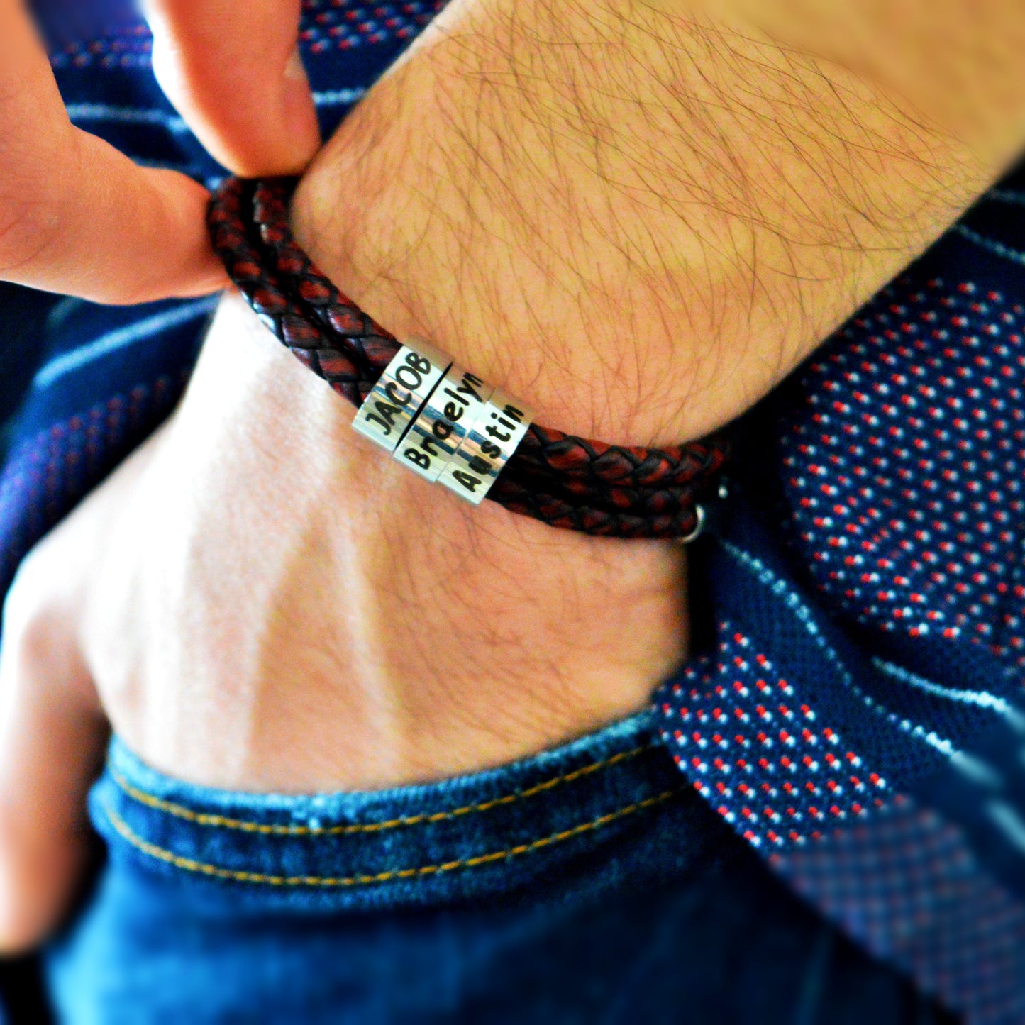 A close-up of a person's wrist wearing a dark brown braided cord bracelet with a silver-colored metal combination lock tag engraved with some names.