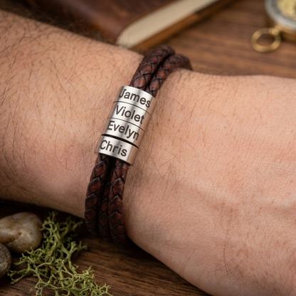 A close-up of a person's wrist wearing a dark brown braided cord bracelet with three engraved silver name plates reading James, Violet, and Evelyn, with another plate reading Chris. The background is a rustic wooden surface with a compass and stones.