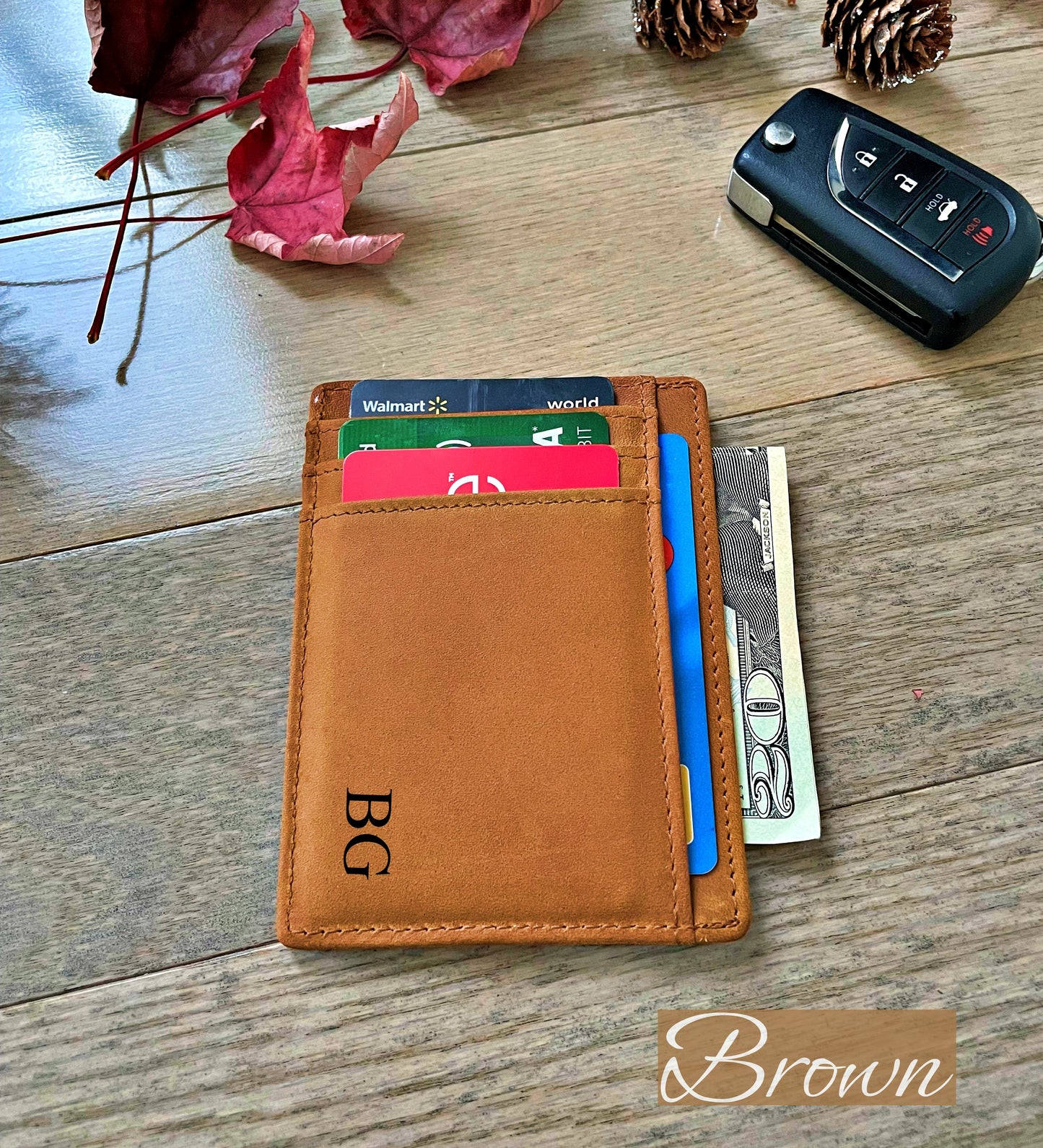 A brown leather minimalist card holder wallet with a monogram 'BG' on the front, shown with cards and cash partially inserted. The wallet is placed on a wooden tabletop next to a car key, with decorative autumn leaves and pinecones in the background.