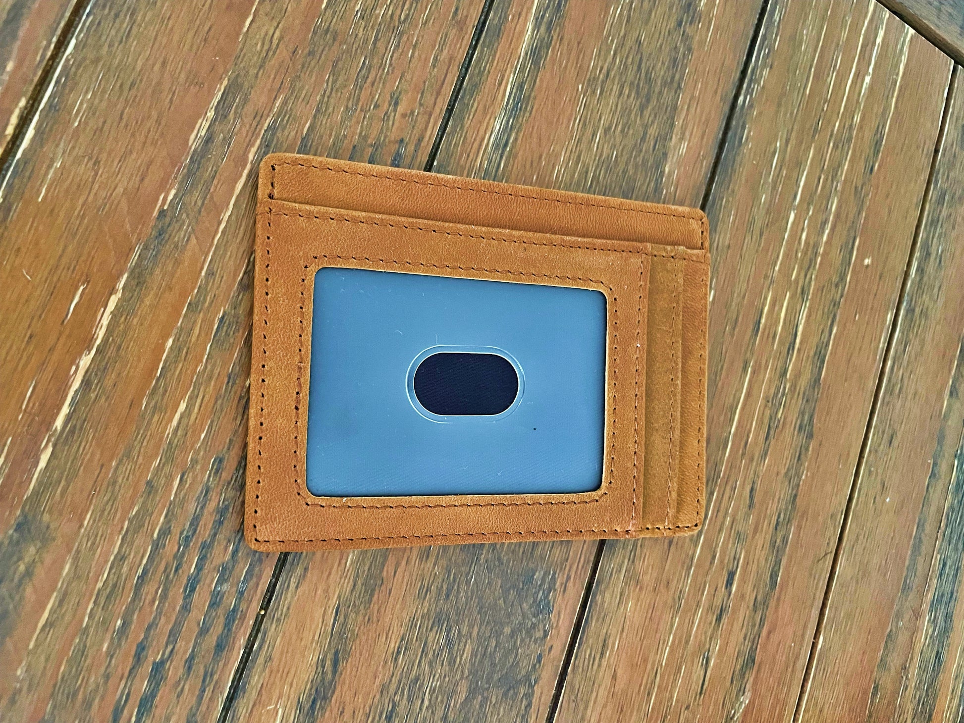 A slim tan leather card holder wallet with a blue ID window, placed on a weathered wooden plank surface.