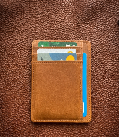 A slim tan leather card holder wallet with multiple card slots and a front slip pocket, shown closed on a brown textured leather background. Several cards are partially visible inside.