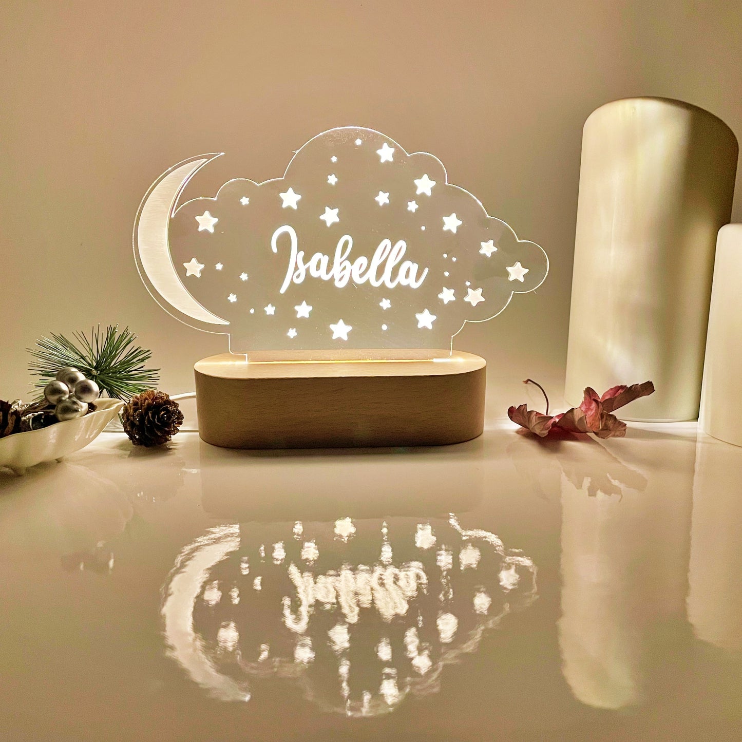 A personalized cloud-shaped night light with a crescent moon and stars, featuring the name 'Isabella' in script, displayed on a wooden base in a warm, cozy setting.