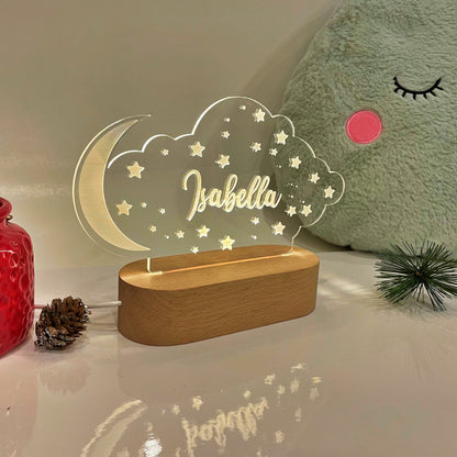 A personalized acrylic night light shaped like a crescent moon and cloud with the name 'Isabella' in gold script, illuminated on a wooden base. The lamp is styled on a glossy white surface next to a red candle holder, pinecones, and a large green plush pillow.