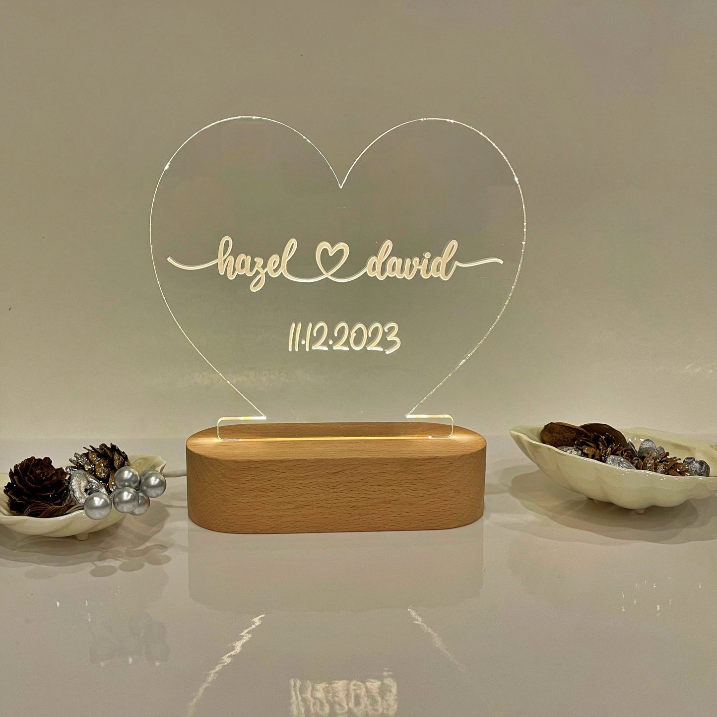 A personalized acrylic heart-shaped light on a wooden base, displaying the names 'hazel david' and the date '11.2.