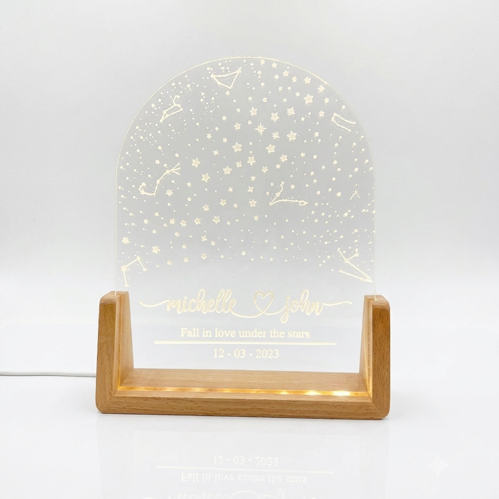 A personalized acrylic night light with a clear arched panel featuring engraved stars, constellations, and custom names and date, displayed on a light wood stand with warm white LED lighting on a white background.