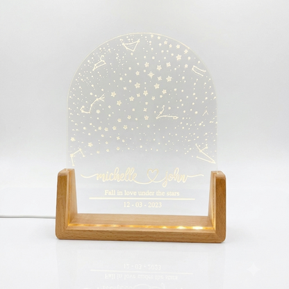A personalized acrylic night light with a clear arched panel featuring engraved stars, constellations, and custom names and date, displayed on a light wood stand with warm white LED lighting on a white background.