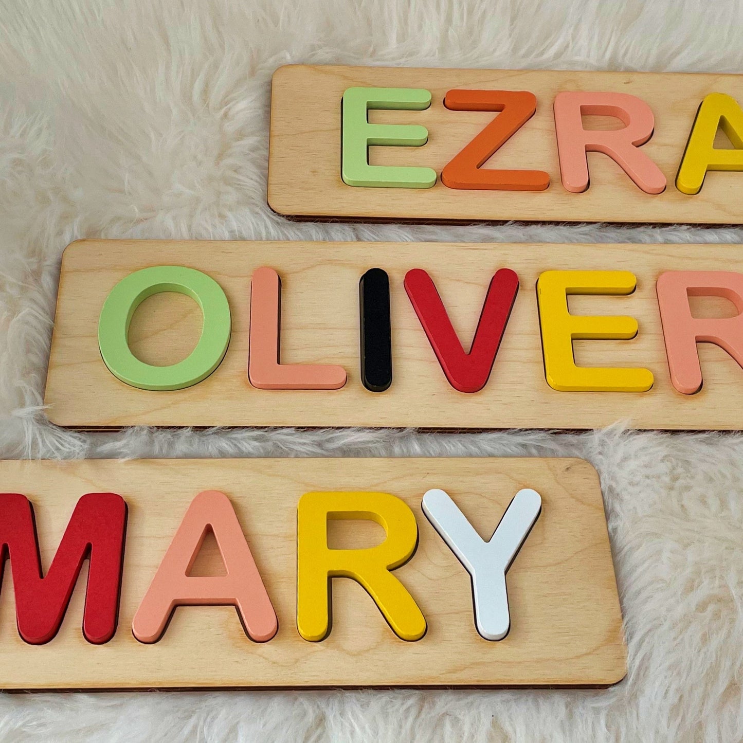 Three wooden nameplaques spelling EZRA, OLIVER, and MARY, each with colorful raised letters on a soft white faux-fur background.