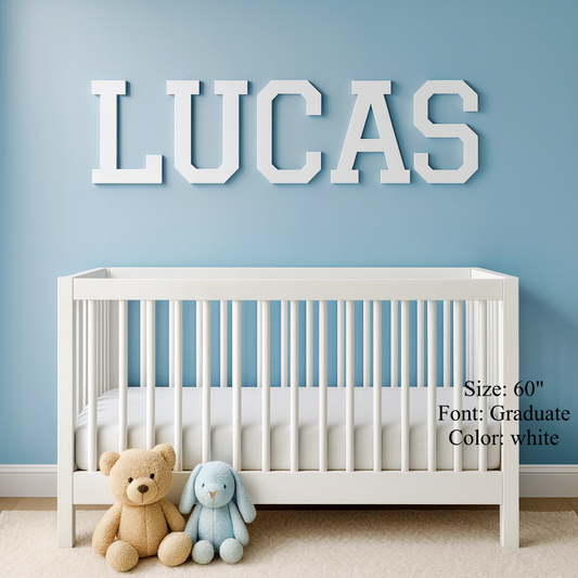 Large white block letters spelling 'LUCAS' mounted on a light blue wall above a white crib, with teddy bears at the foot of the crib. Text on the right reads 'Size: 60" Font: Graduate Color: white'.